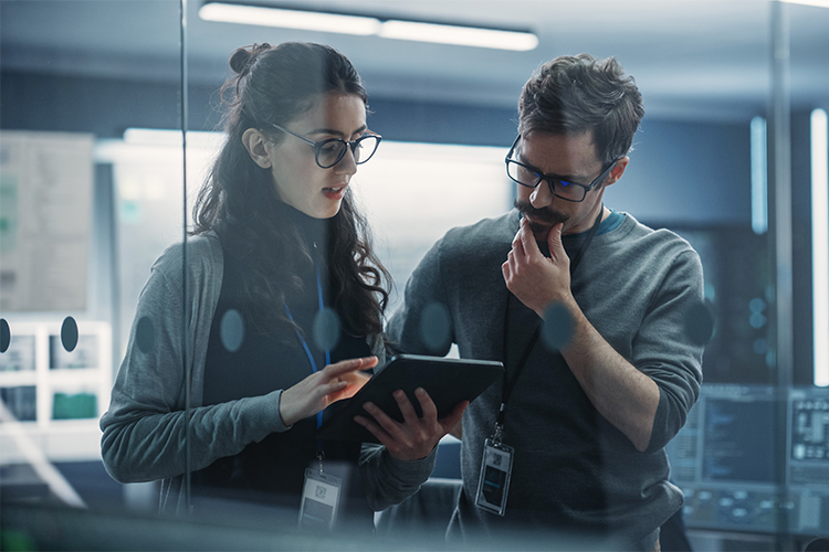 Retrato de dois jovens engenheiros criativos, uma mulher e um homem, usando um tablet para analisar e discutir como proceder com o software de inteligência artificial. Eles estão em pé em um escritório de pesquisa de alta tecnologia.