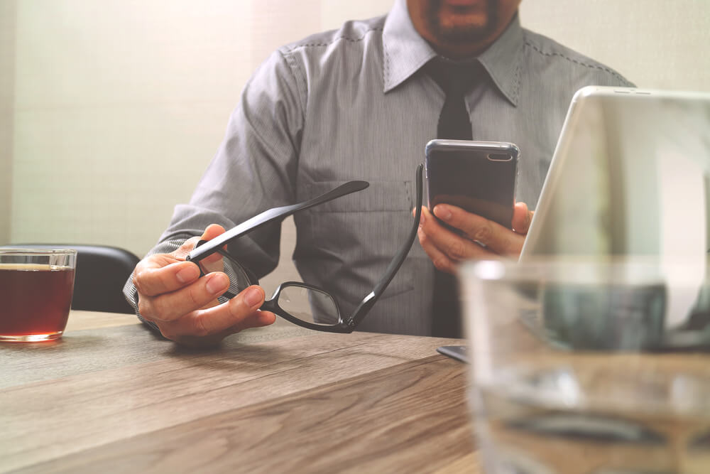 Homem com óculos na mão usando celular na outra