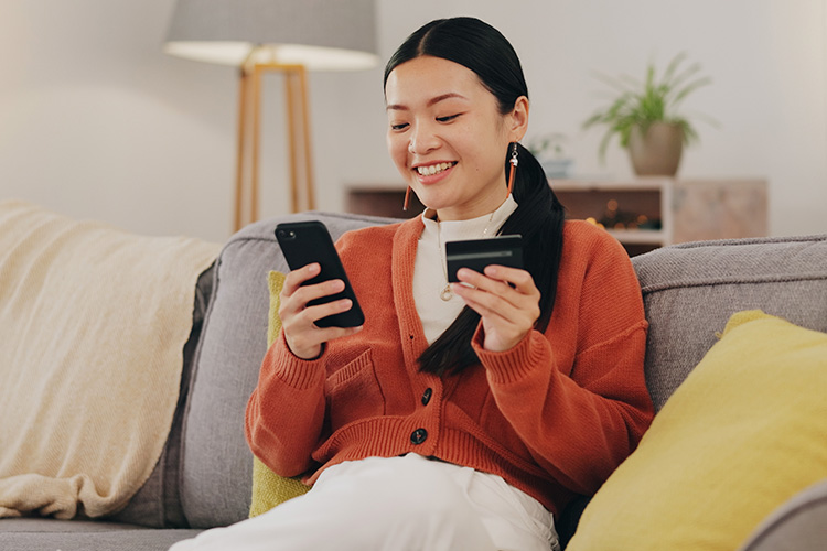 Uma mulher feliz está sentada no sofá, utilizando seu celular e cartão de crédito para fazer compras online, utilizando facilidades de pagamento oferecidas pelo comércio eletrônico e tecnologia financeira.